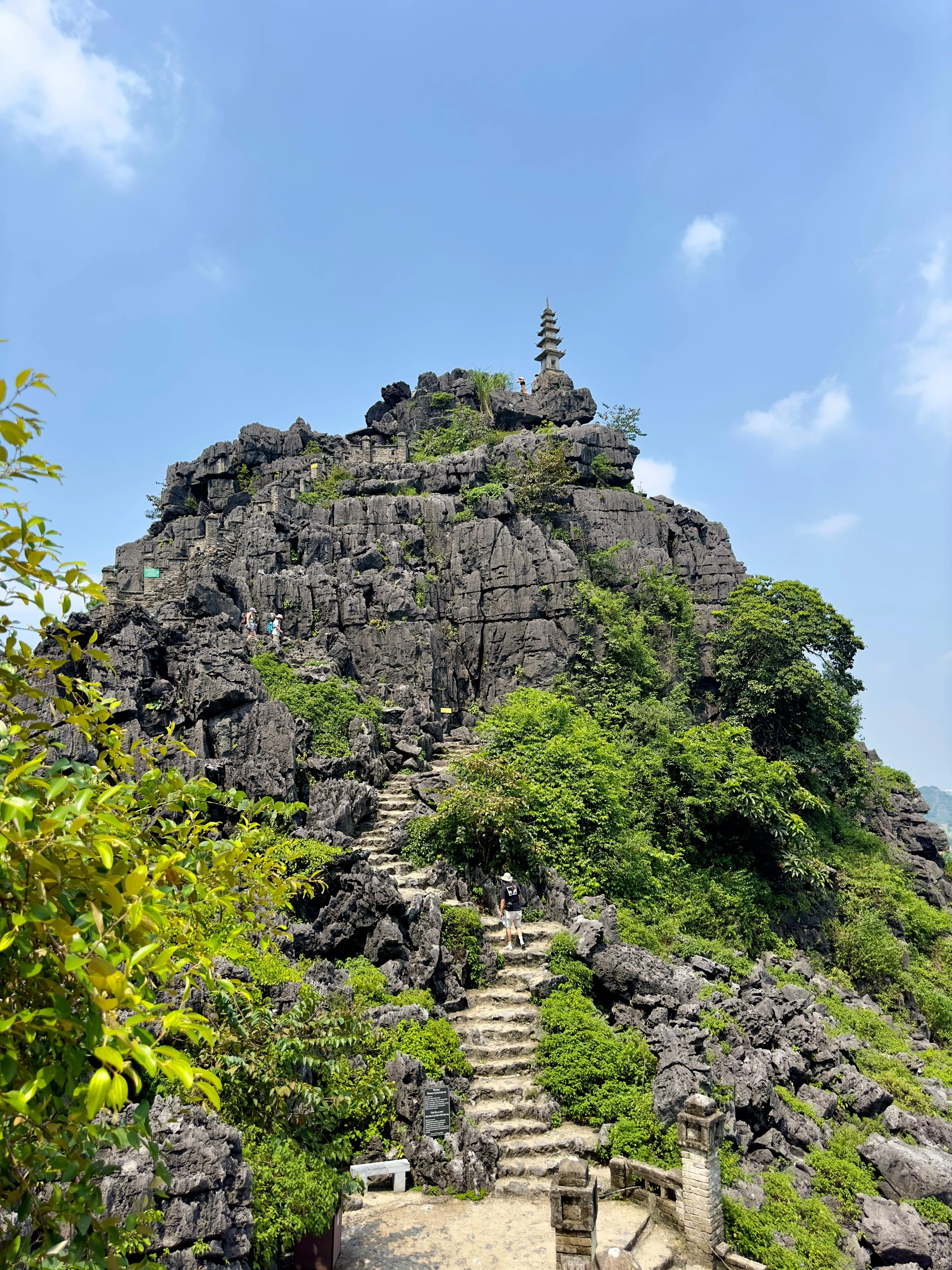Een stenen trap leidt naar een berg met een pagode op de top, omgeven door groene struiken en rotsen, onder een blauwe hemel met enkele wolken.