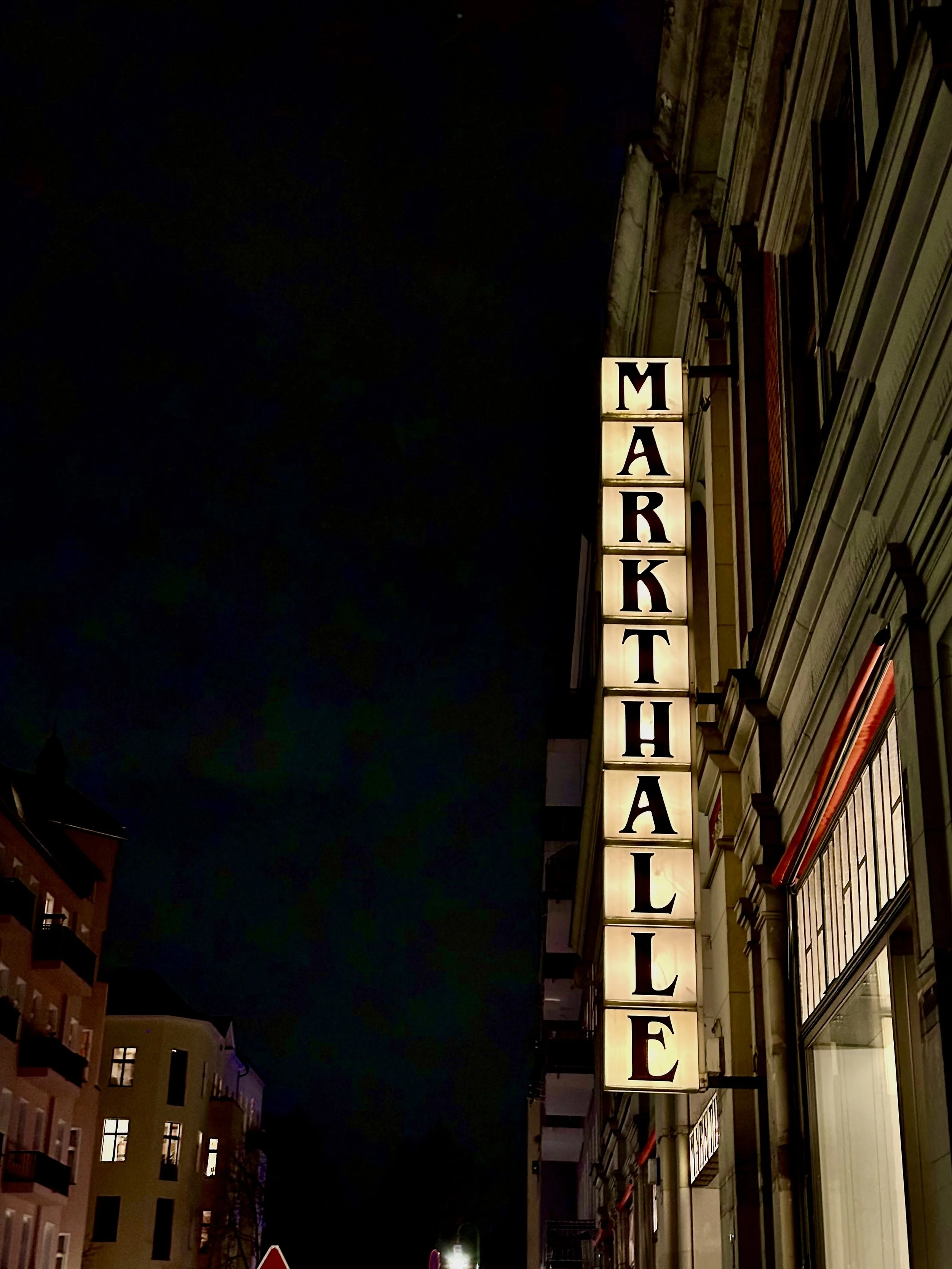 Verlichte stadszicht met een verticale neonbord met de tekst 'MARKTHALLE' op een gebouw bij nacht.