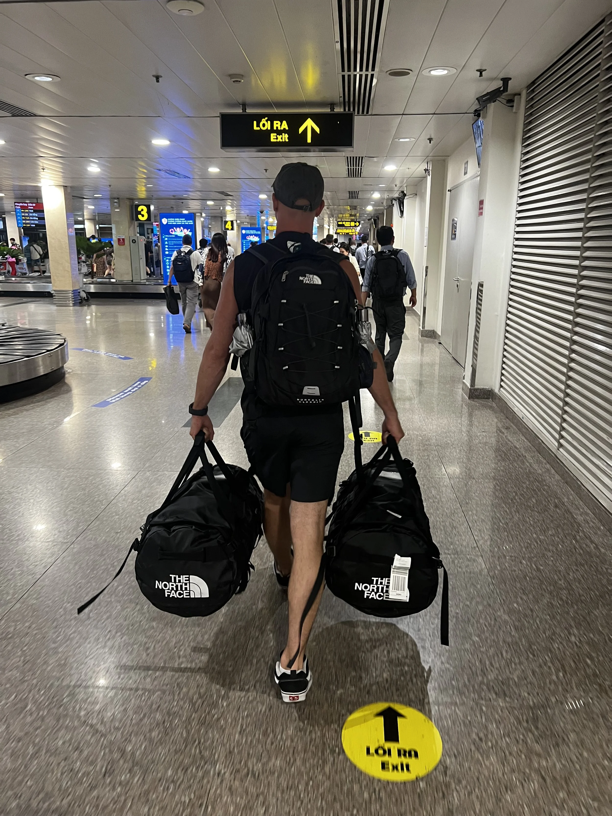 Man met een backpack en twee reistas loopt door een luchthaven. Er hangen digitale informatieschermen en bewegwijzering, en andere passagiers lopen voor hem.