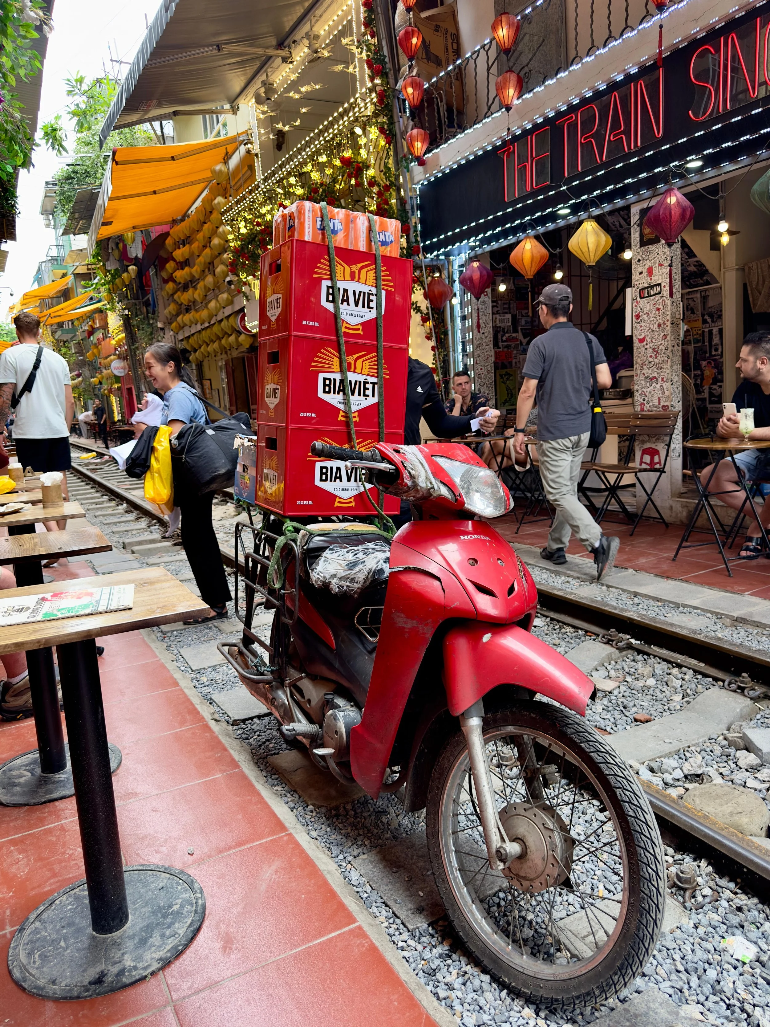 Trainstreet in Hanoi. Een straat met een rode scooter geparkeerd naast een terras, omringd door mensen die wandelen en zitten. De straat is versierd met gele en rode lampionnen en lichte decoraties.
