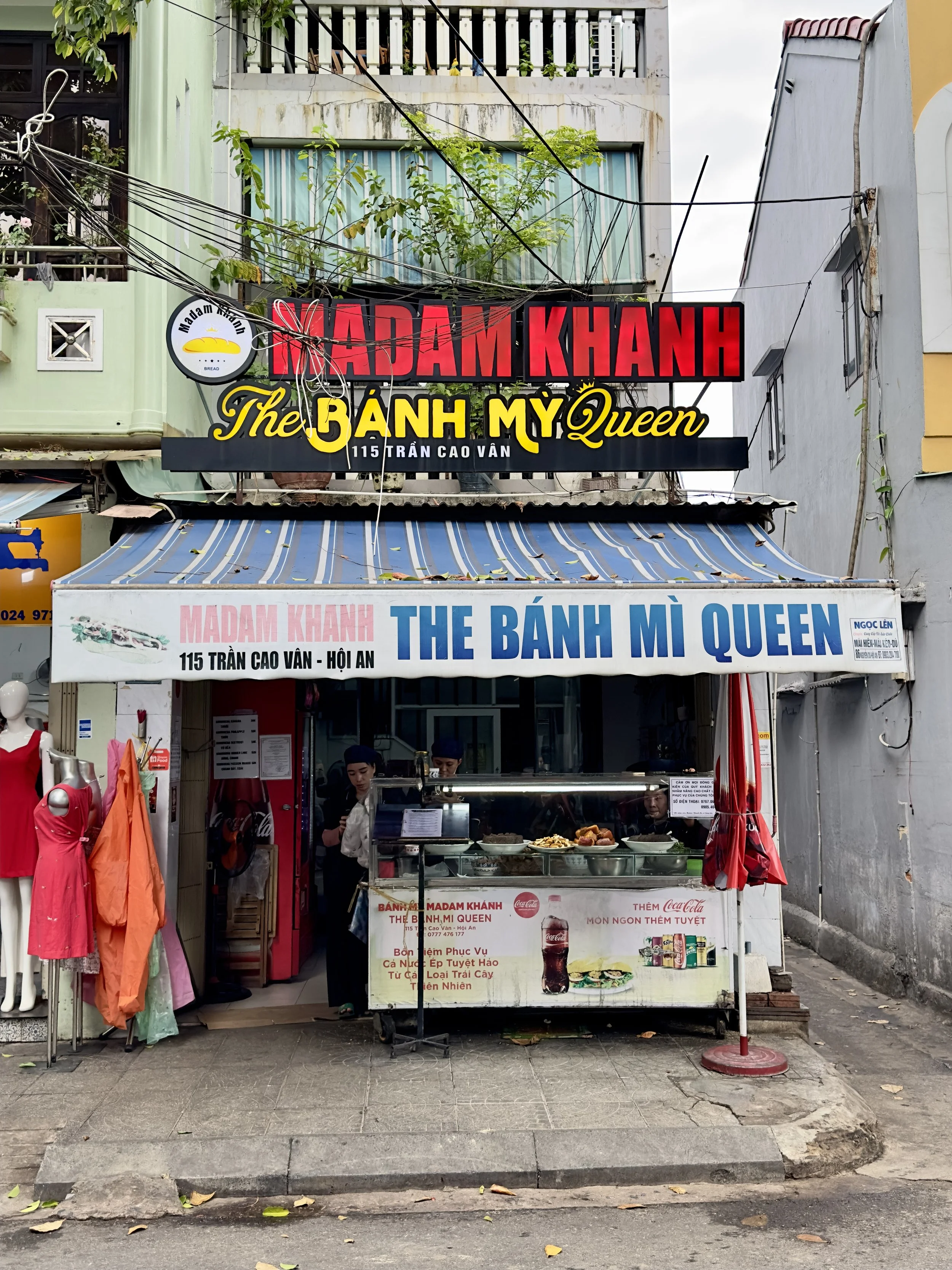 Een straatkraam dat Vietnamese borden of sandwiches verkoopt, met mensen die eten bereiden en klanten die wachten. Verschillende reclameborden en een winkelcase met eten en drankjes.