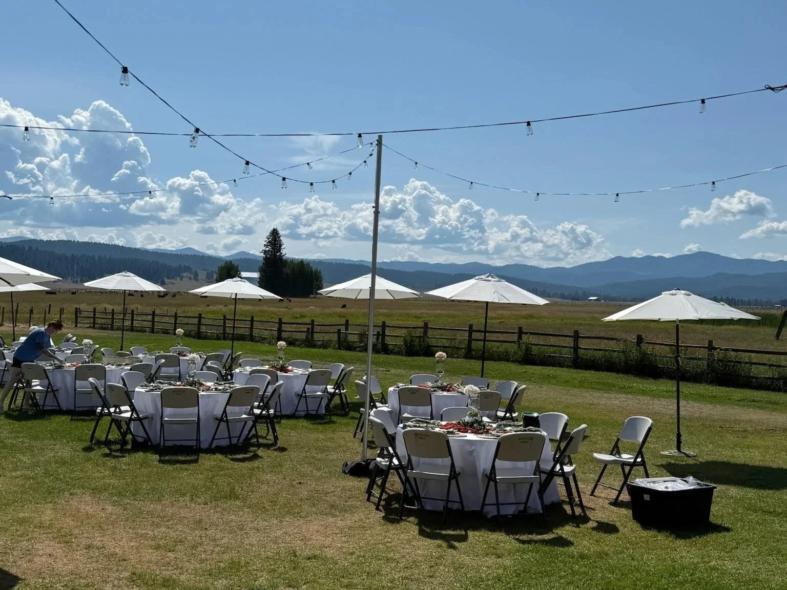 Event setup for an outdoor wedding with umbrellas, tables, chairs, and bistro lighting