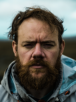 Close-up of Richard Glover with a beard and disheveled hair, wearing a blue jacket outdoors.