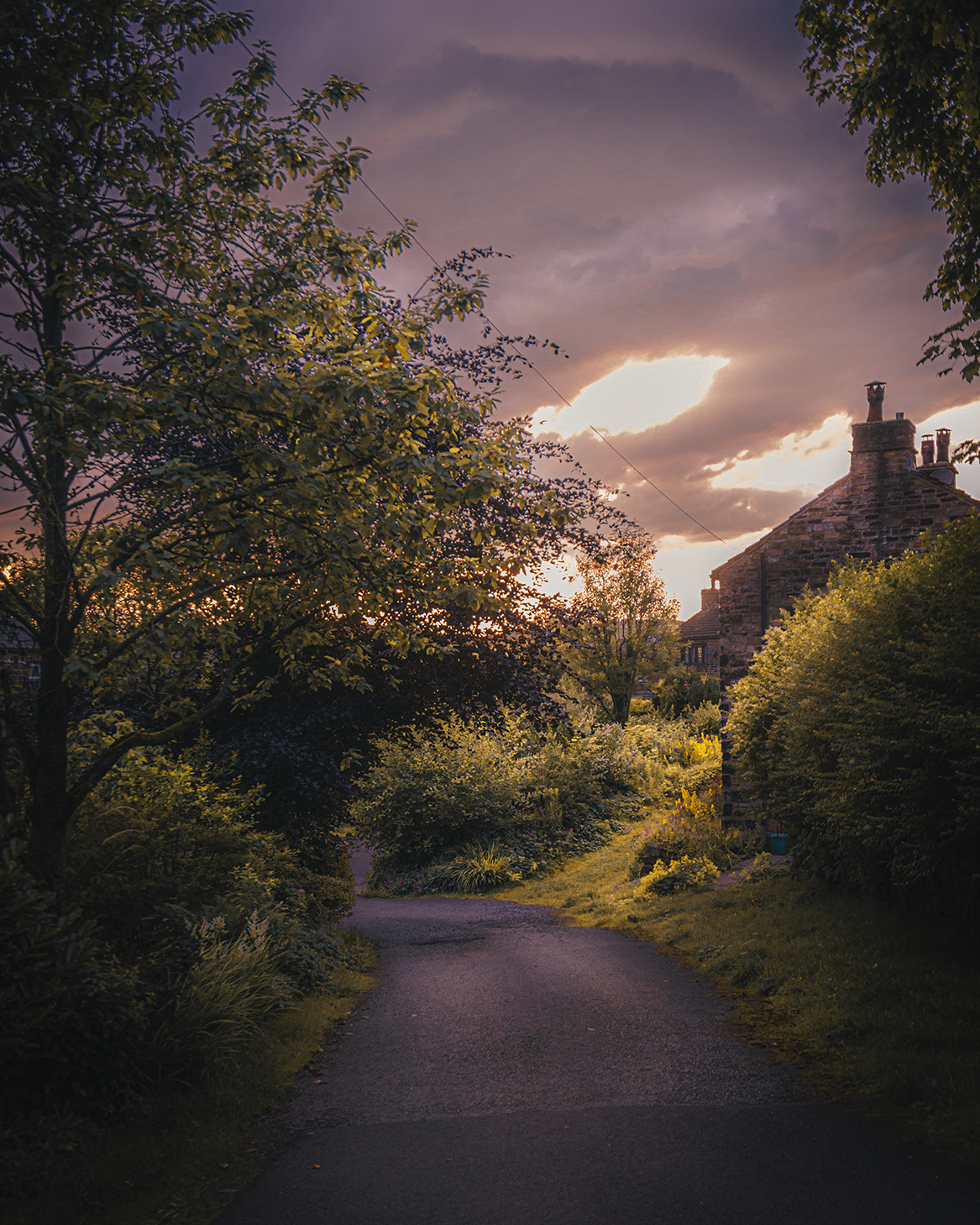 A winding country road surrounded by lush green trees and bushes, with a stone house on the right and a sky with dark clouds and a partially visible setting sun in the background.