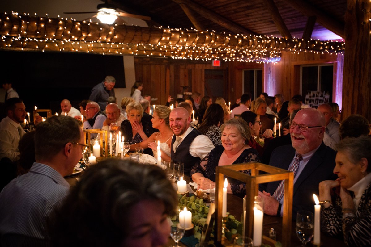 Reception by candlelight in a lodge. View looking down the table as bride and groom smile and laugh with guests.