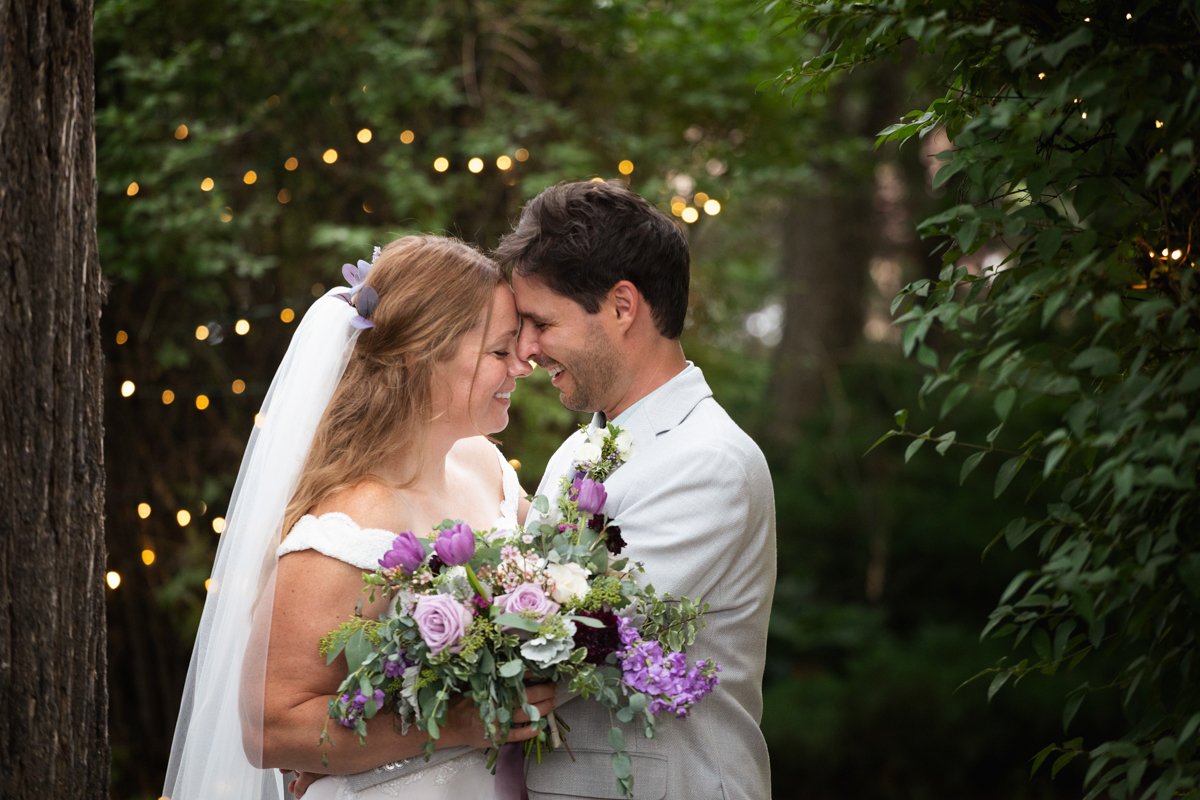 A couple embracing while holding a purple flower bouquet in a thicket of bushes with twinkle lights behind them.