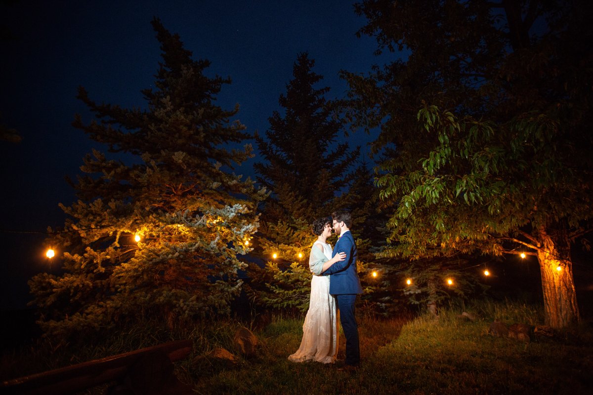 Couple embrace at night in a grove of pine trees with cafe lights in them.