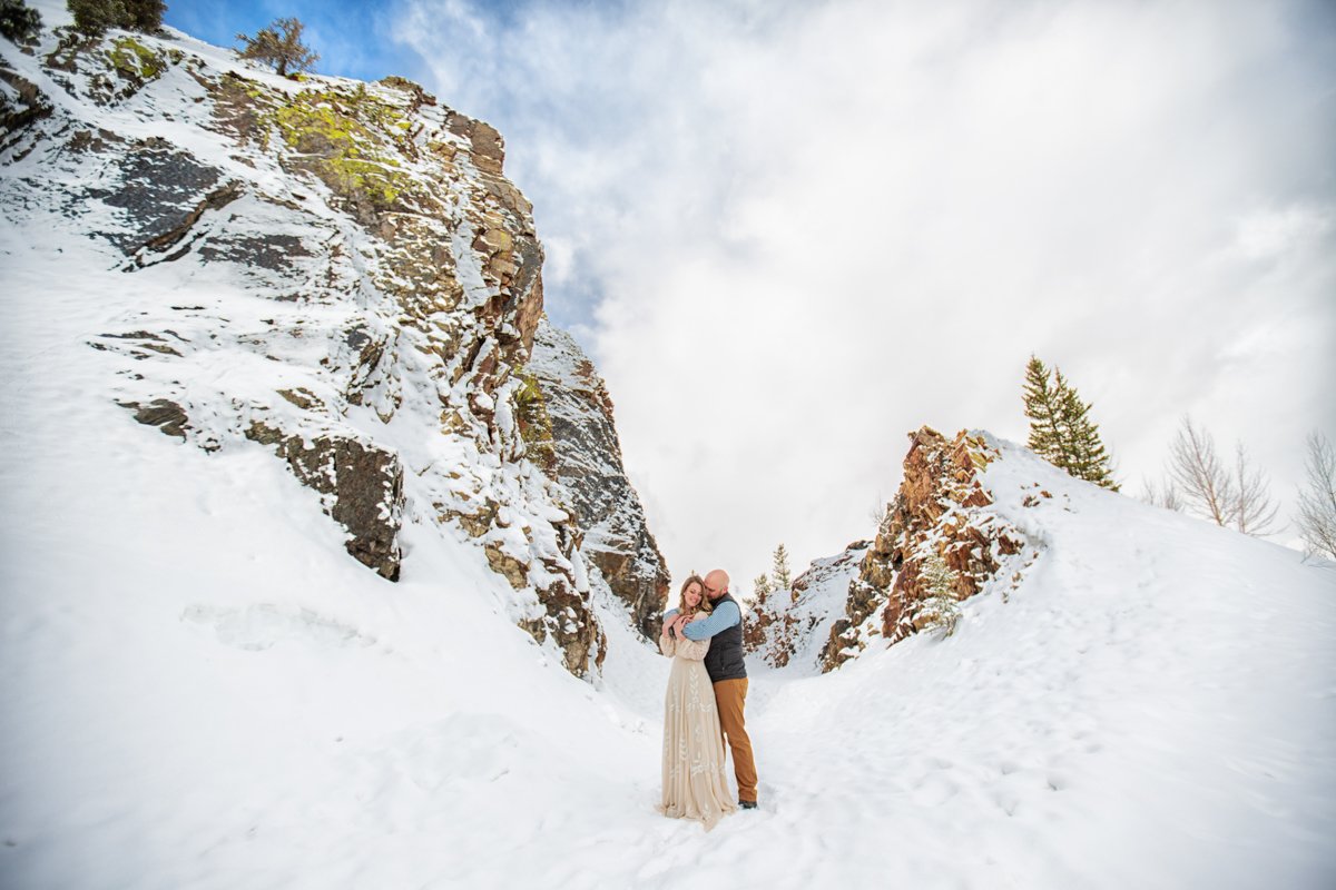 A man hugs a woman from behind while they stand in the snow in front of a large rock formation.