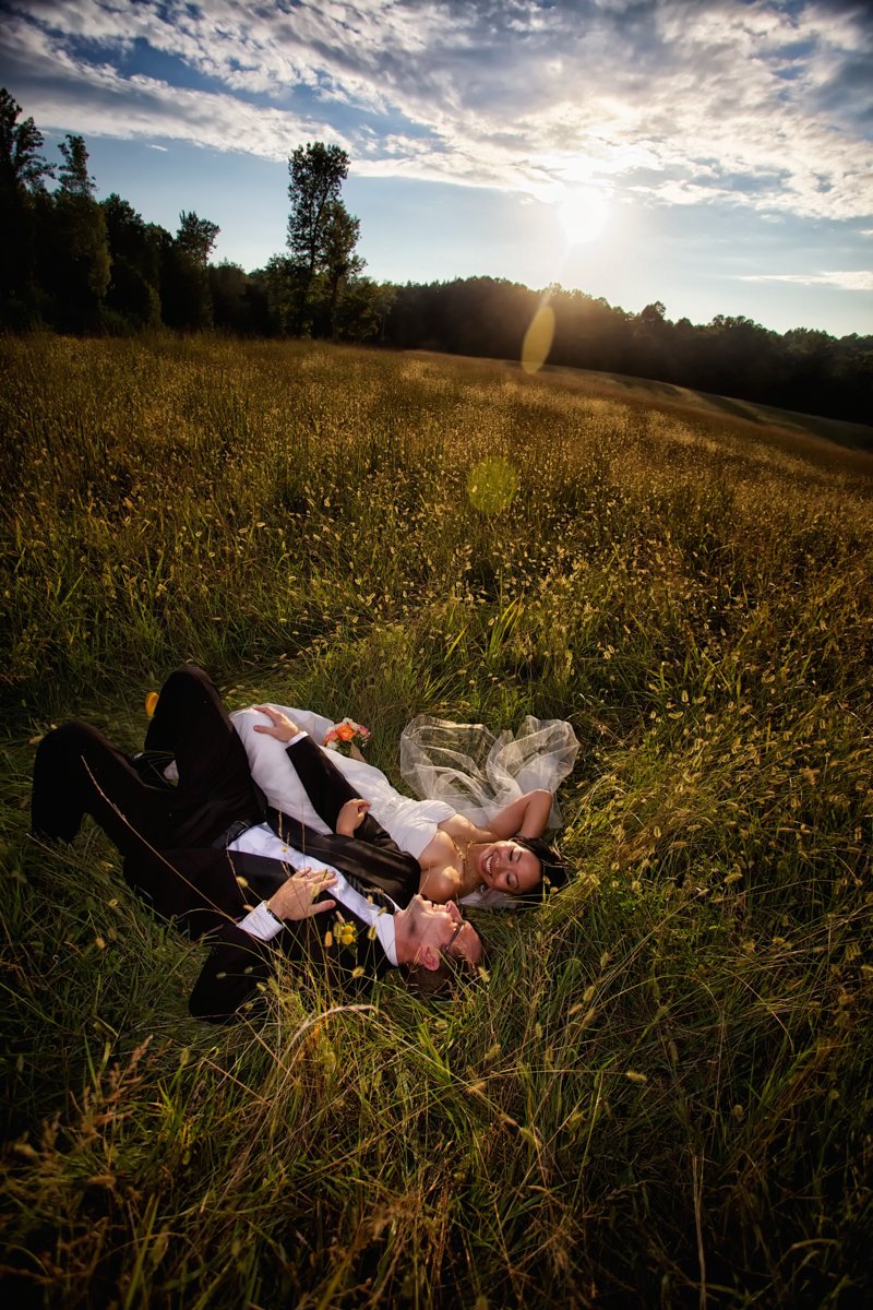 A couple lays in a field, side by side, hand in hand, at sunset. The sun offers a lens flair and catches the light of the tops of the grass that has gone to seed. Romantic and whimsical.