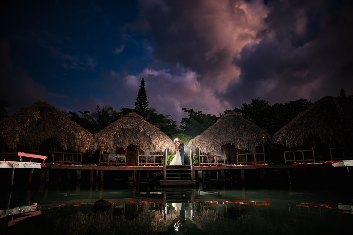 Couple in Belize standing on a dock with grass huts behind them. They're illuminated and the sky is dark and blue and purple with giant clouds. They are reflected in the water.