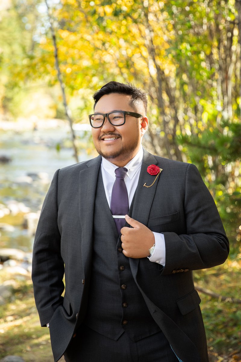 Groom stands on the shore of a river at fall.