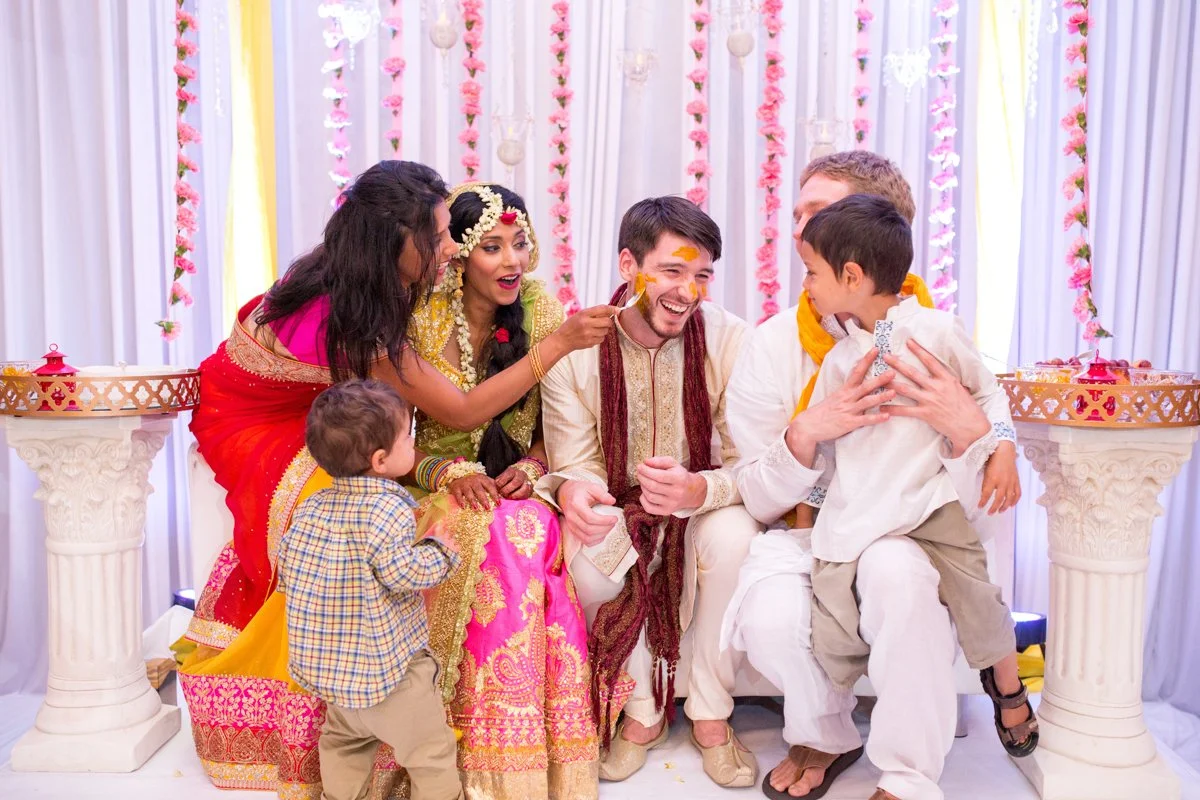 Bride's family paints groom's face with turmeric paste during Indian wedding ceremony. They sit on a stage with garlands of flowers behind them.