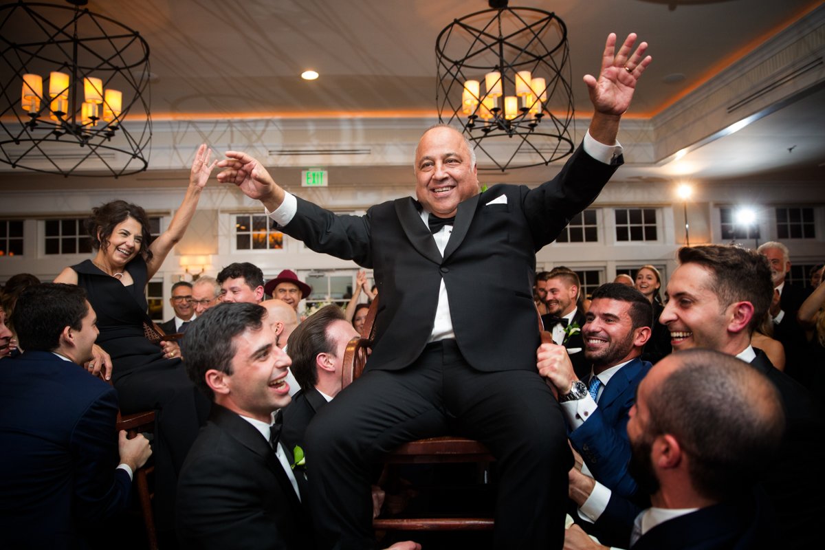 Parents of the bride raised high on chairs in a hora during a wedding reception. Laughing, with their hands in the air.