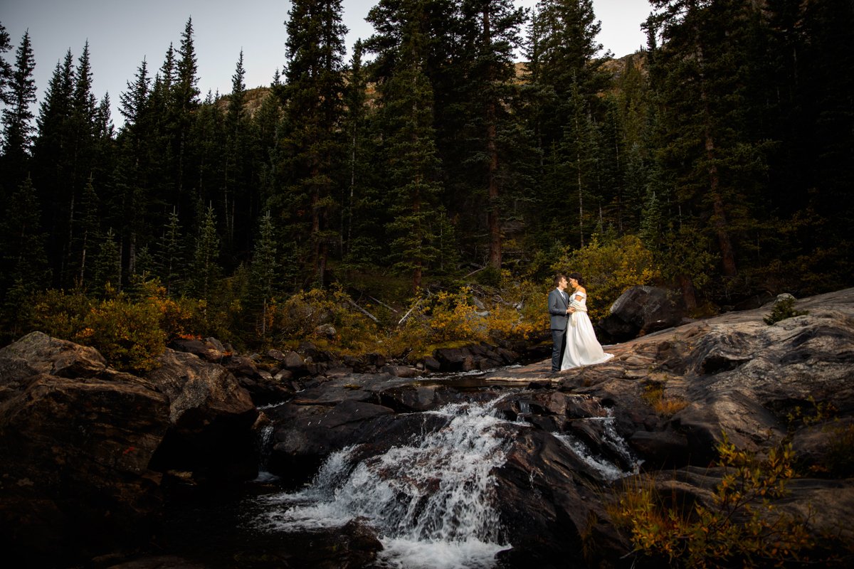 Couple on a rock ledge beside a waterfall in their wedding attire surrounded by tall, dark, evergreens.