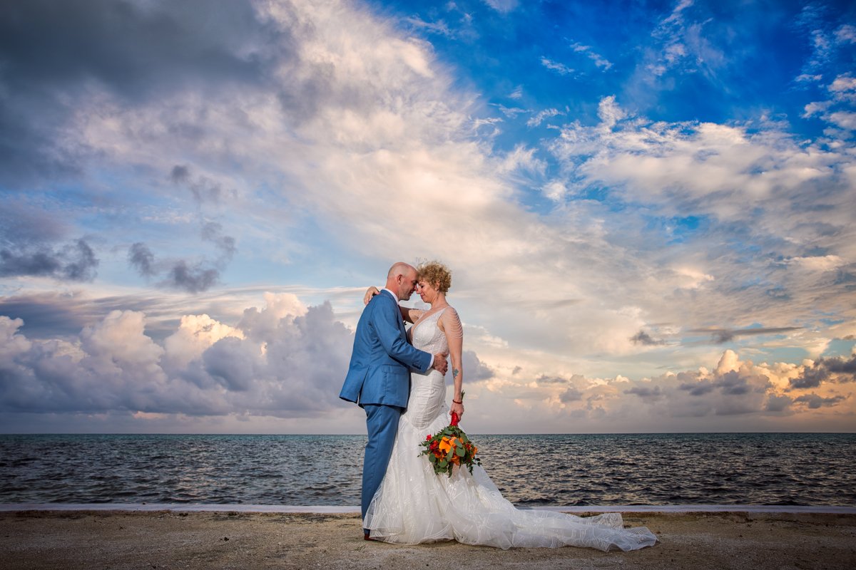 Couple embraces on the beach in Belize. with dramatic blue clouds. Groom wears blue. Bride wears a trumpet wedding dress with tropical flower bouquet.