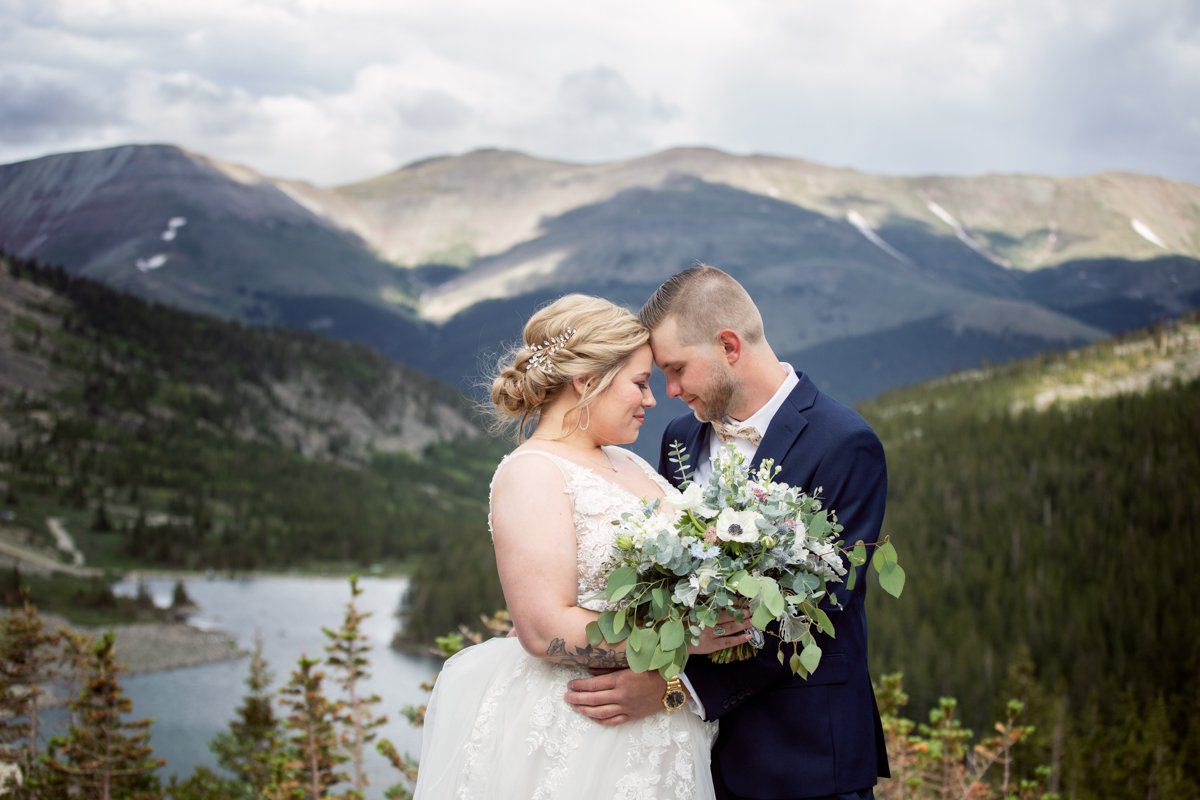 Couple stands on a cliffside, up high, forehead to forehead while holding a bouquet. A lake and mountains are behind.