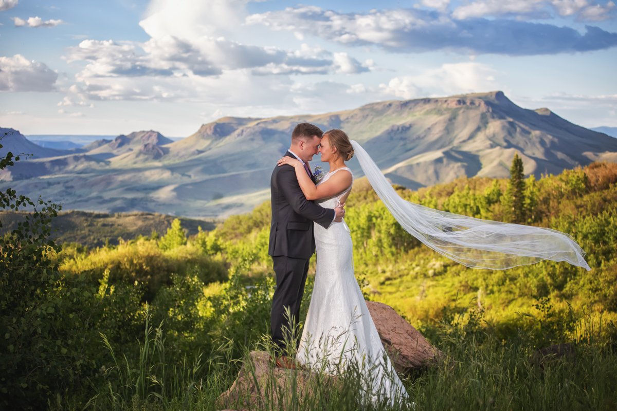 Couple embrace, face to face, while standing on a rock overlooking a summer mountain waning light. bride's veil trails behind her.