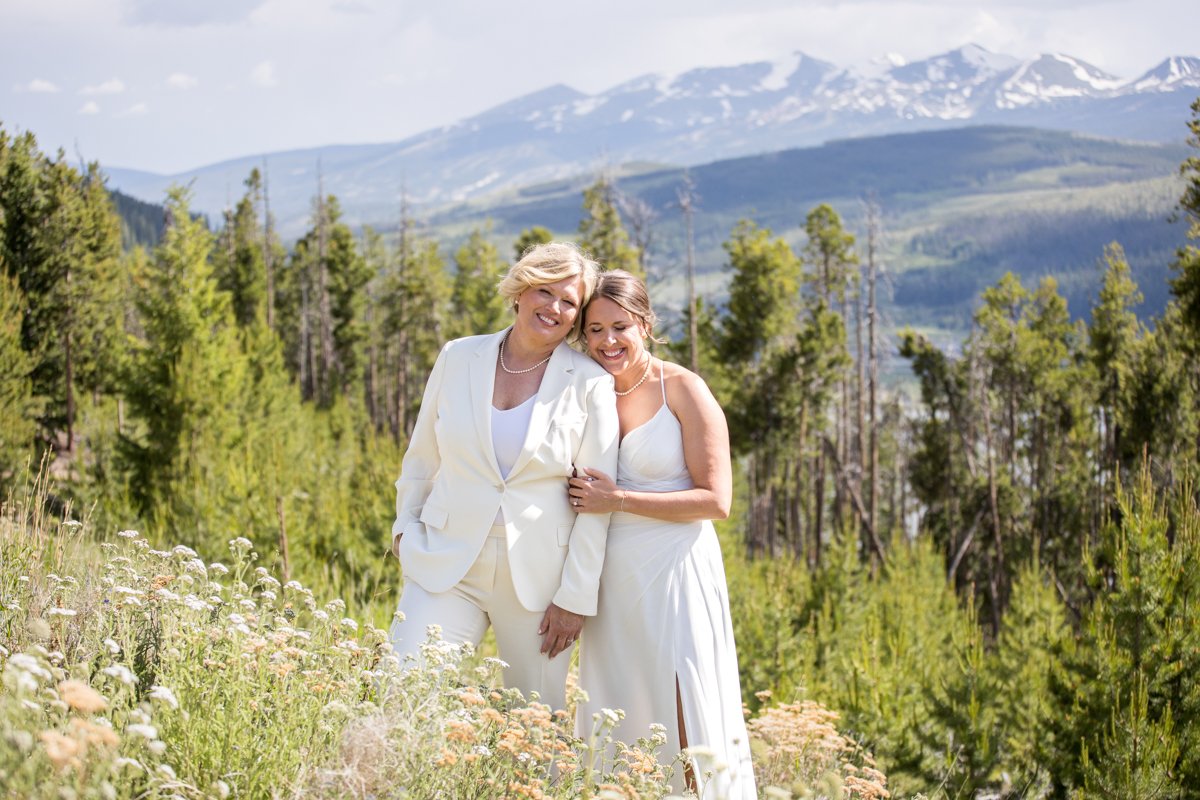 Two brides embrace on a hillside with mountains behind them.