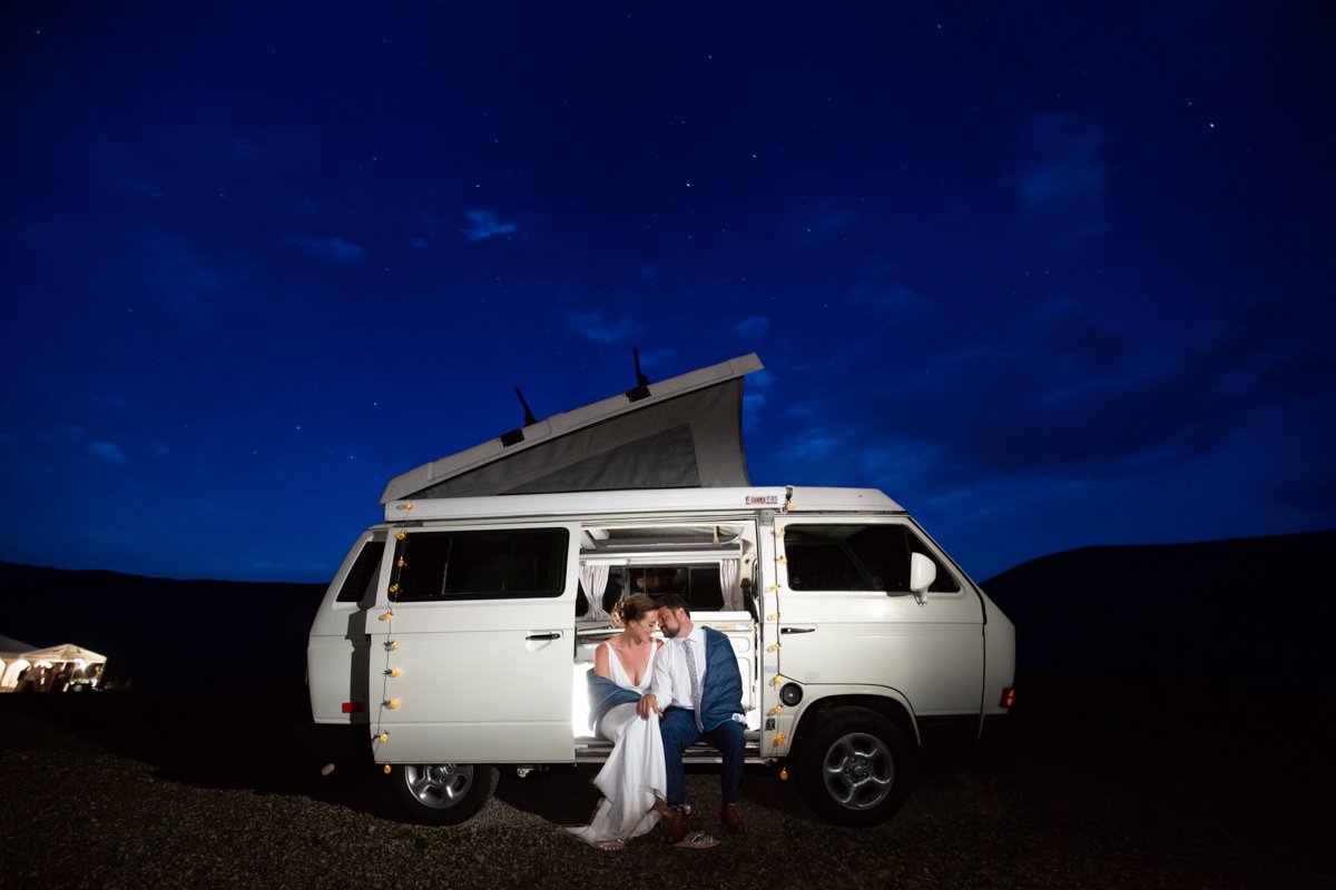 Night portrait of a couple in their camper van. the van is pictured from the side. They're sitting with the sliding side door open and illuminated from the inside. The night sky is deep blue above them. The reception party tent glows in the distance.