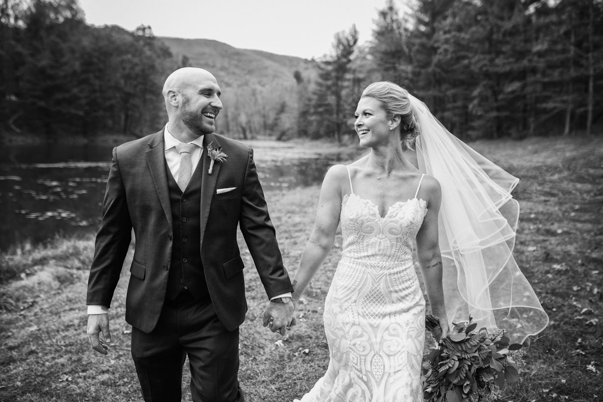 Couple walking hand in hand toward the camera but they look at each other. They're walking in a field next to a lake with mountains.