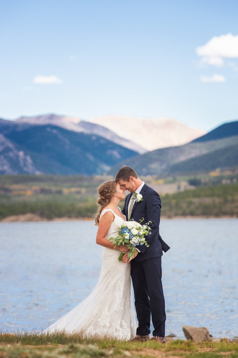 Couple standing lakeside with mountains converging behind them.