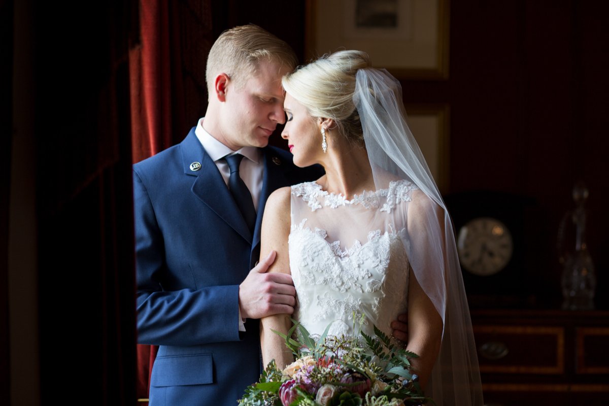 Couple embrace next to window light framed by thick red velvet curtains.