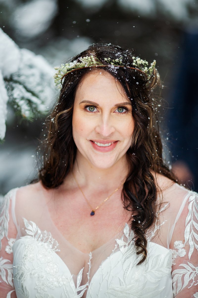 Bridal portrait in the snow. Bride wears an elaborate applique dress overlay and floral crown while snowflakes fall into her hair.