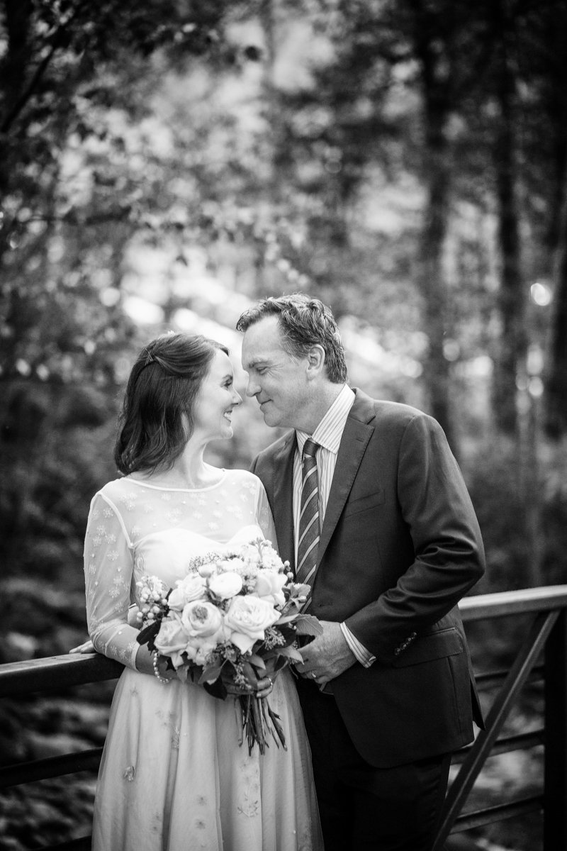 A couple stand on a bridge in the forest. Black and white. They snuggle close to each other.