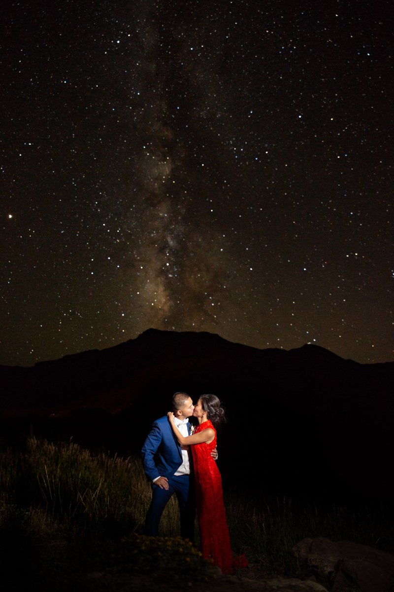 A couple hold each other at night with the milky way visible overhead and the silhouette of a mountain behind them. Groom wears a blue suit. Bride wears a traditional red Chinese gown.