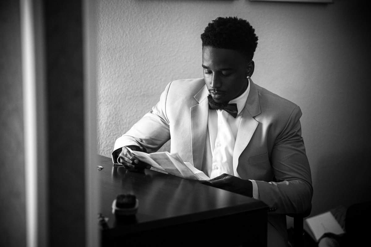 Black and white photo of a groom sitting at a desk reviewing his written vows to his future wife before the ceremony. Photo is taken looking through a doorway.
