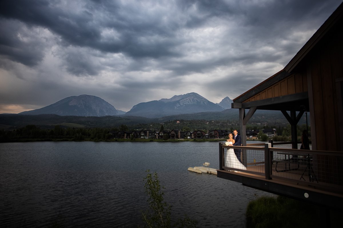 A couple stand on the edge of a deck. The shot is wide and shows the deck overlooking a lake with mountains behind. It's blue hour. The couple is illuminated with lighting from the photographer.