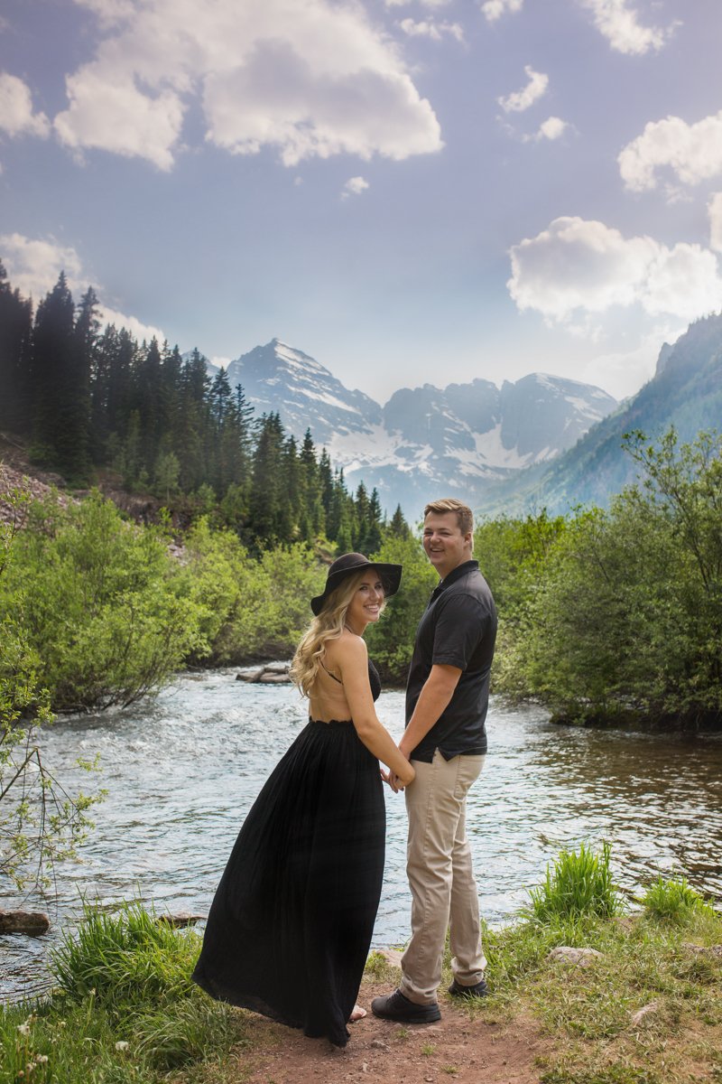 Couple standing hand in hand looking back at the camera, on a lake shore overlooking Maroon Bells.