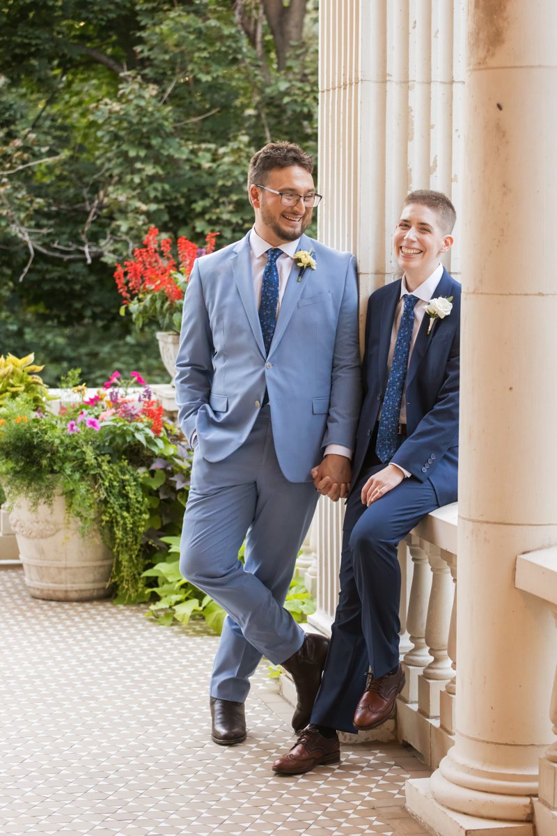 Two grooms at their ceremony location just after their ceremony. One sits on a railing while the other leans against a column. They hold hands.