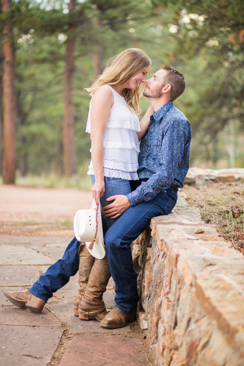 Engagement photo for a couple. Man sitting on a stone wall, woman standing in front of him, about to kiss. She's holding his cowboy hat.