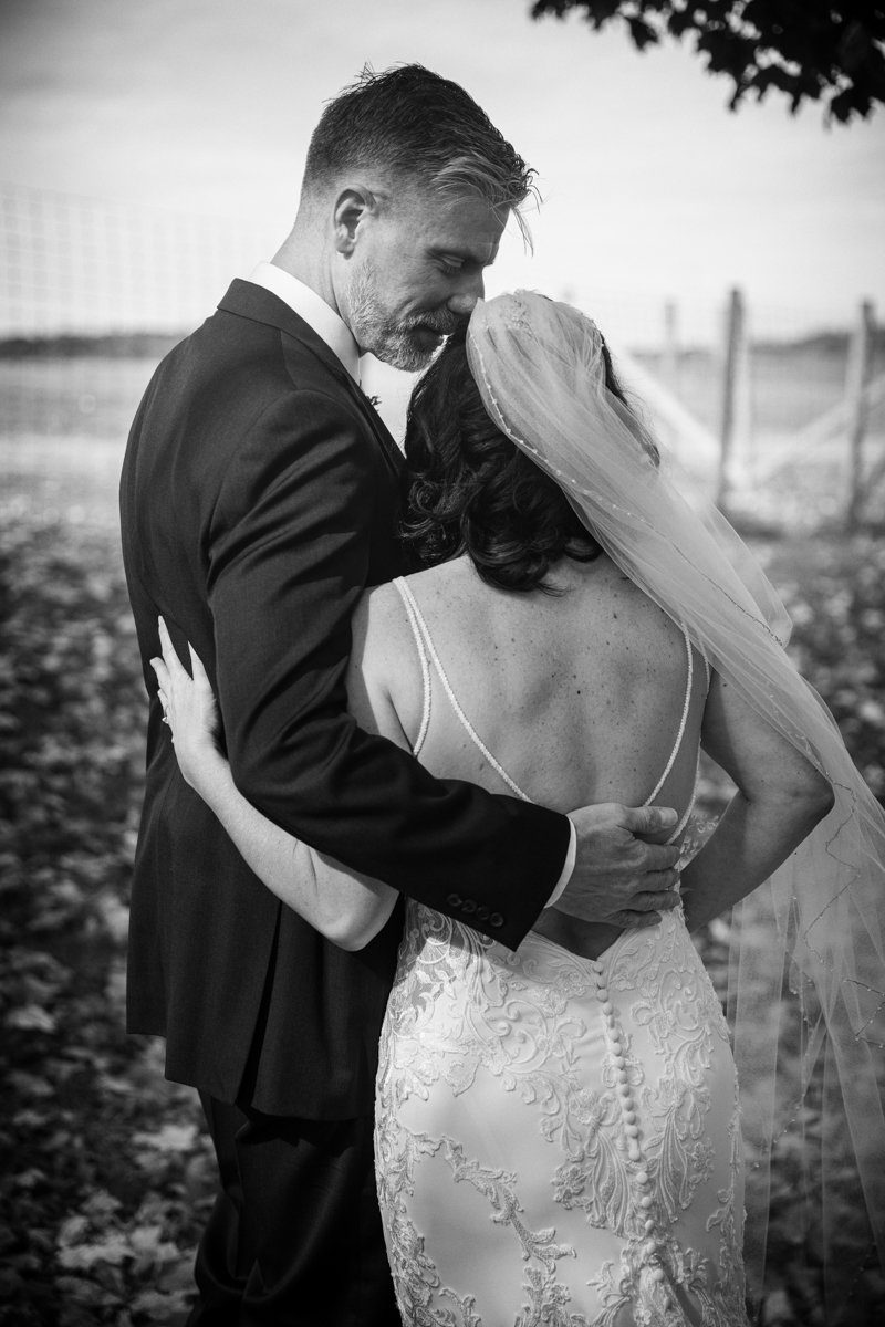 Quiet moment in black and white of a very tall groom embracing his bride. Shows off the back of her dress with embroidery and small buttons.