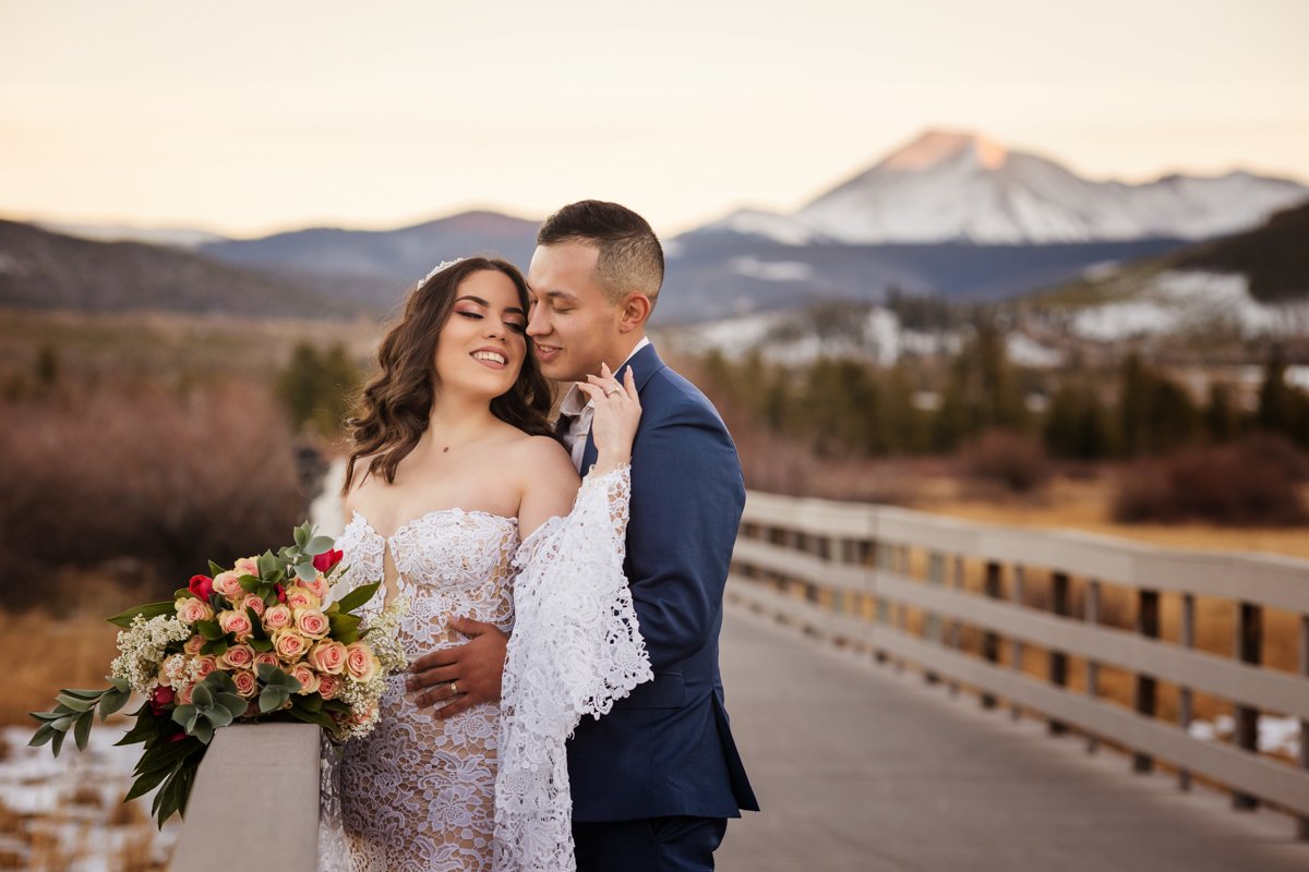 A couple at dusk on a bridge with a snow capped mountain behind them. The bride ears a lace gown with long lace sleeves. She faces the walls of the bridge with her flowers on the railing. She turns toward her groom who snuggles up behind her.