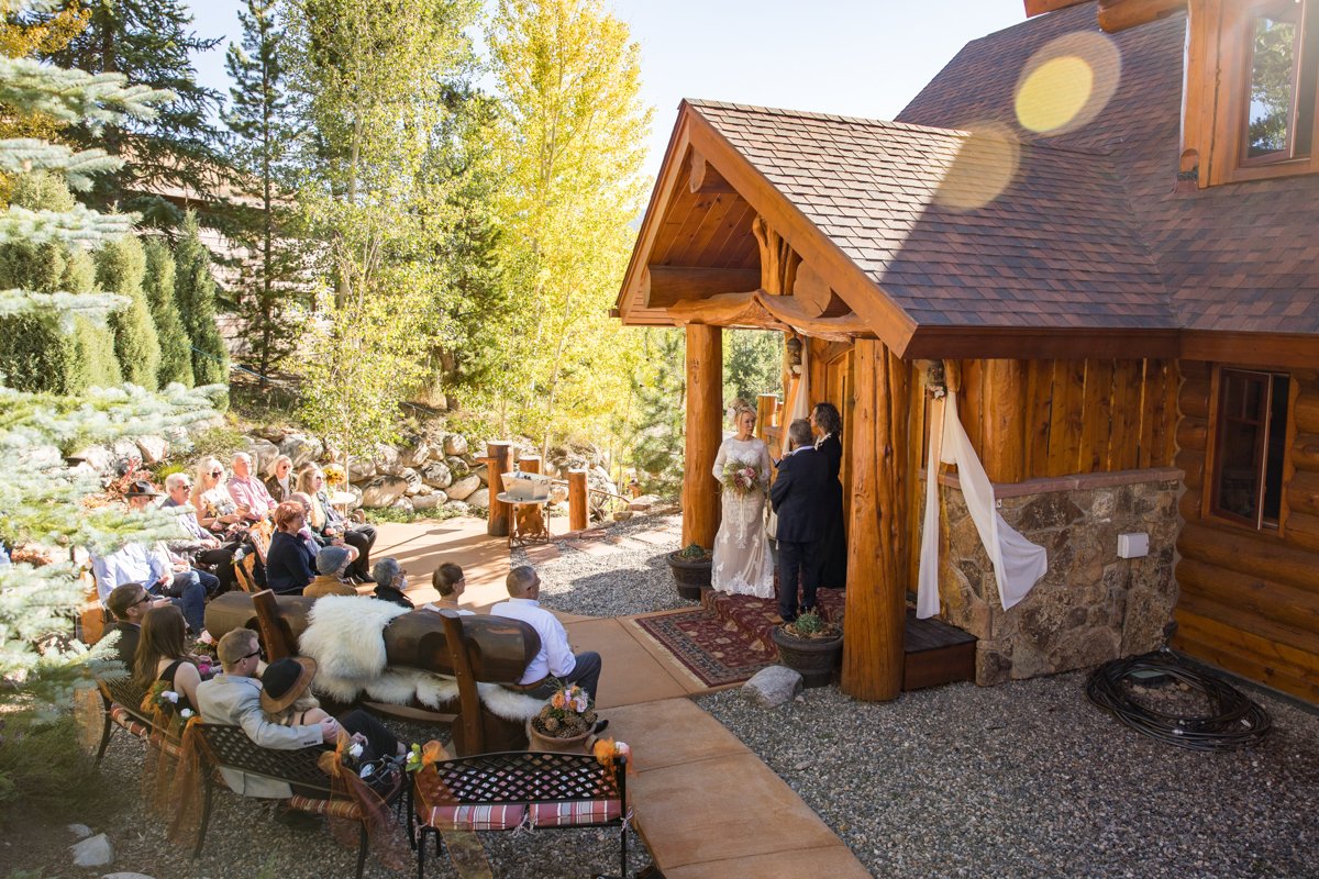 Couple weds at their mountain home front porch with friends and family seated and watching. Log cabin home. Forest around them.