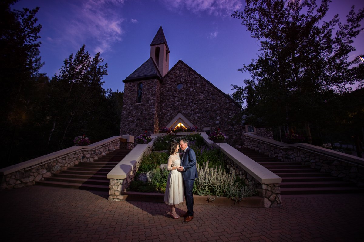 Couple standing in front of the Beaver Creek Chapel at sunset. The sky is pink and blue and purple and the couple is illuminated