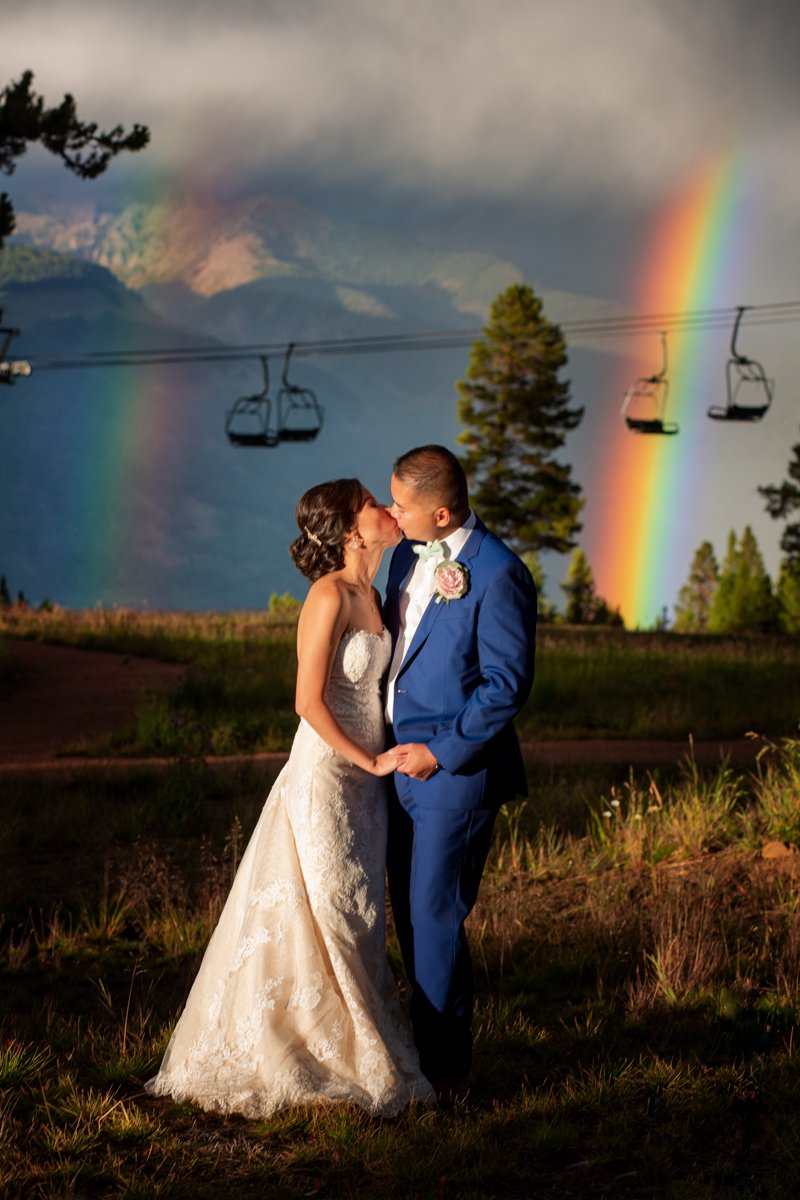Couple kiss in front of ski lifts with a double rainbow and mountains behind them at Vail Ski Resort.