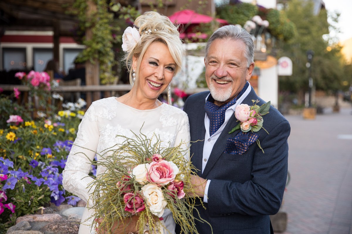 Mature couple jubilantly smiling after their nuptials. Bride wears a white gown with long sleeves and has a whimsical bouquet of greenery and pink roses. They pose in front of wildflowers in Vail Village.