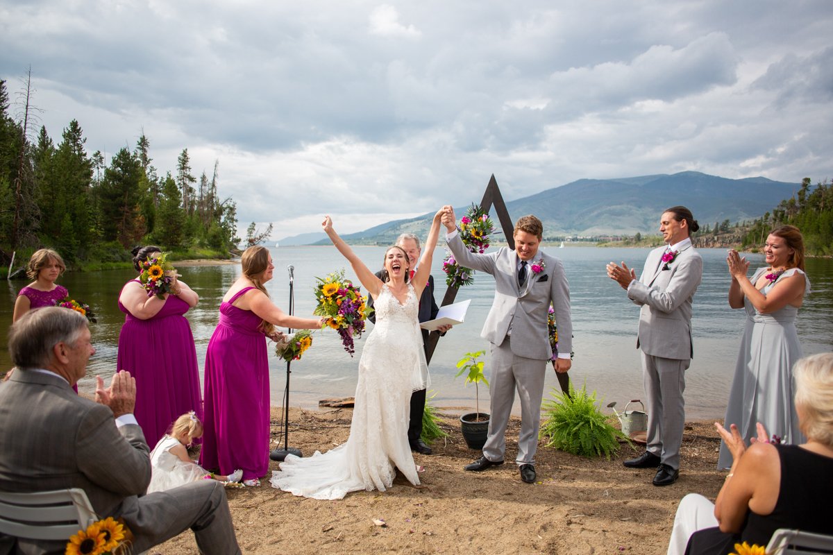 Couple cheers after their vows on a beach of an alpine lake with mountains in the background.