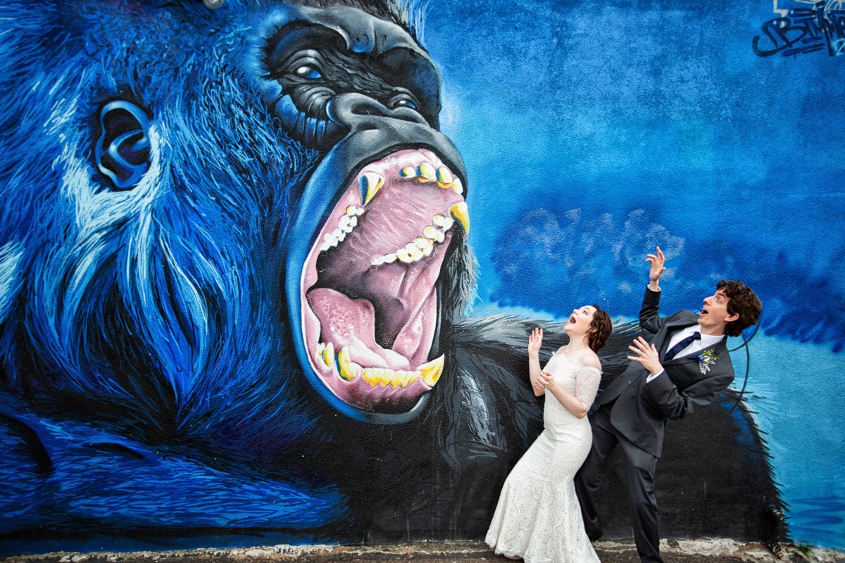 A couple get playful with a street art mural of godzilla roaring.