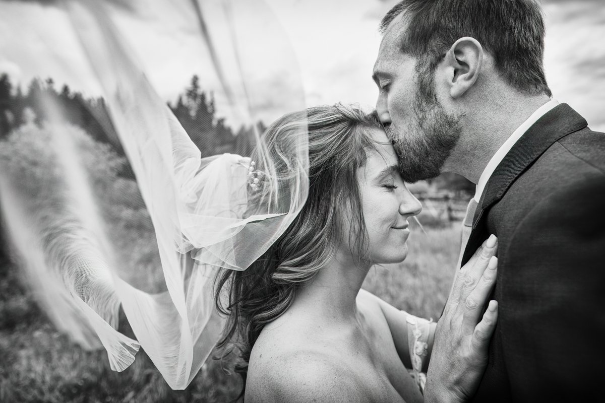 Couple stands facing each other. Groom kisses bride on the forehead. Both their eyes are closed. Her veil is caught in the wind and blowing toward the camera.