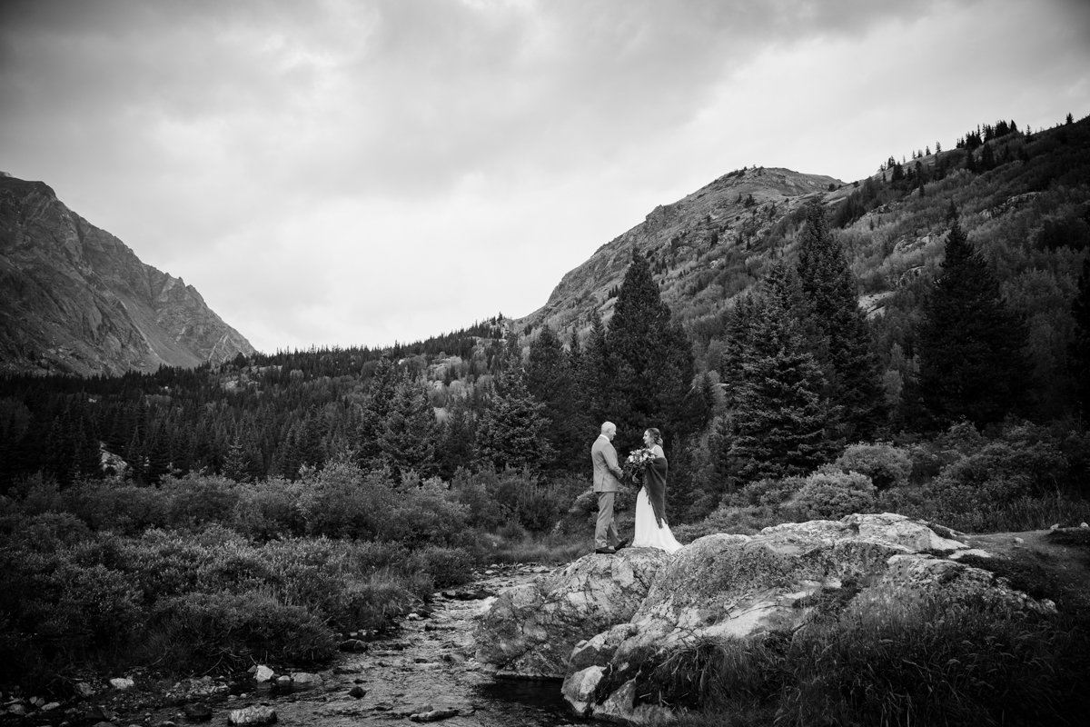 Couple standing on a boulder beside a creek in the mountains as they recite vows to each other. Image is black and white.