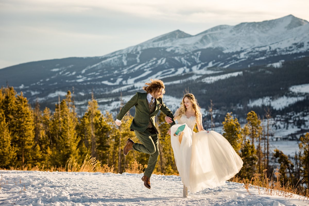 A couple leaps for joy--mid air--on a snow covered path. Breckenridge ski resort ski slopes are snow covered behind them. Sunset.