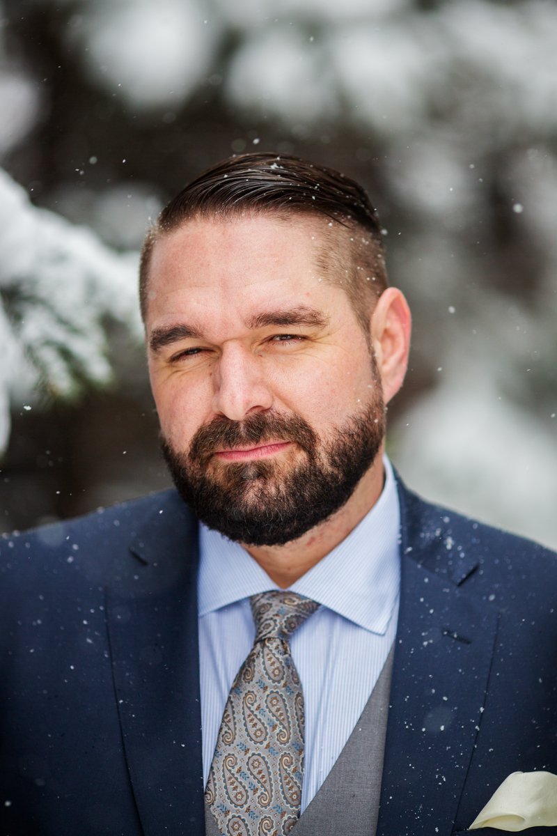 Groom portrait in the snow. He looks at the camera with a dashing expression as snow falls into his hair and around him.