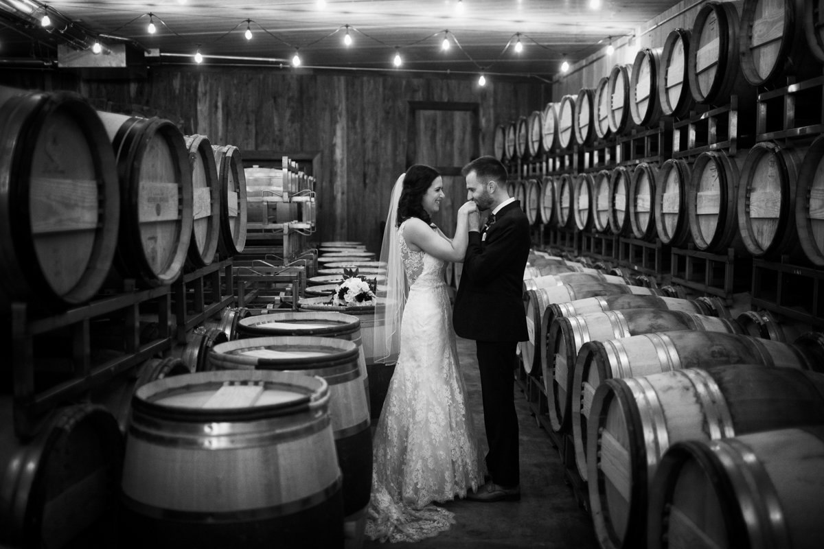 Black and white photo of a couple facing each other. The groom kisses the bride on the hand. They stand in a barrel room of a winery.