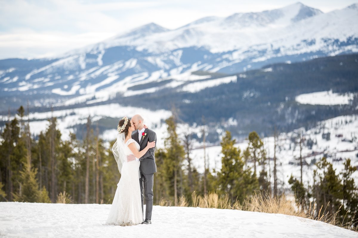 A couple embrace and kiss on a snow covered path with ski slopes and snow covered mountains behind them.