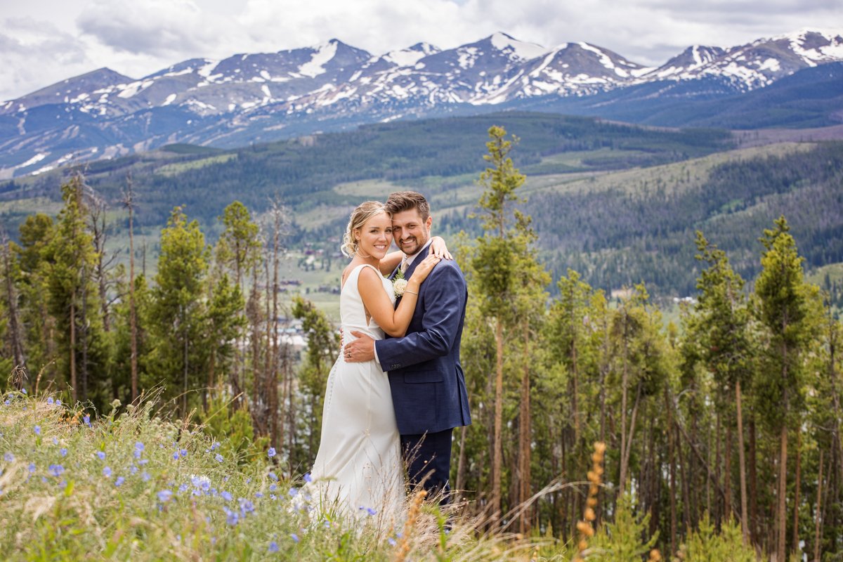 Couple embrace on a hillside with snowcapped mountains behind them.