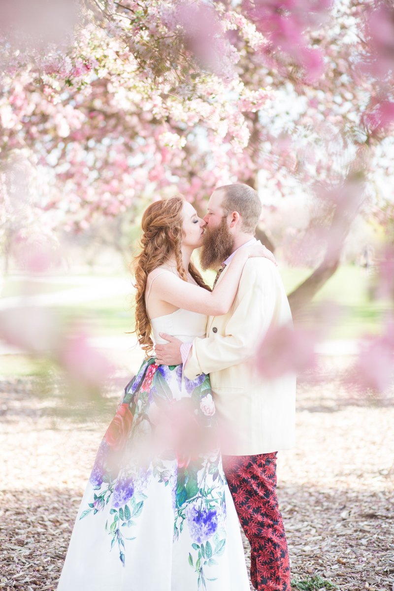 Artsy wedding couple kissing under pink blossoming trees with blossoms layered in the scene. Bride wears a white dress with blue, purple, and green florals. Groom wears a cream jacket with red floral pants.